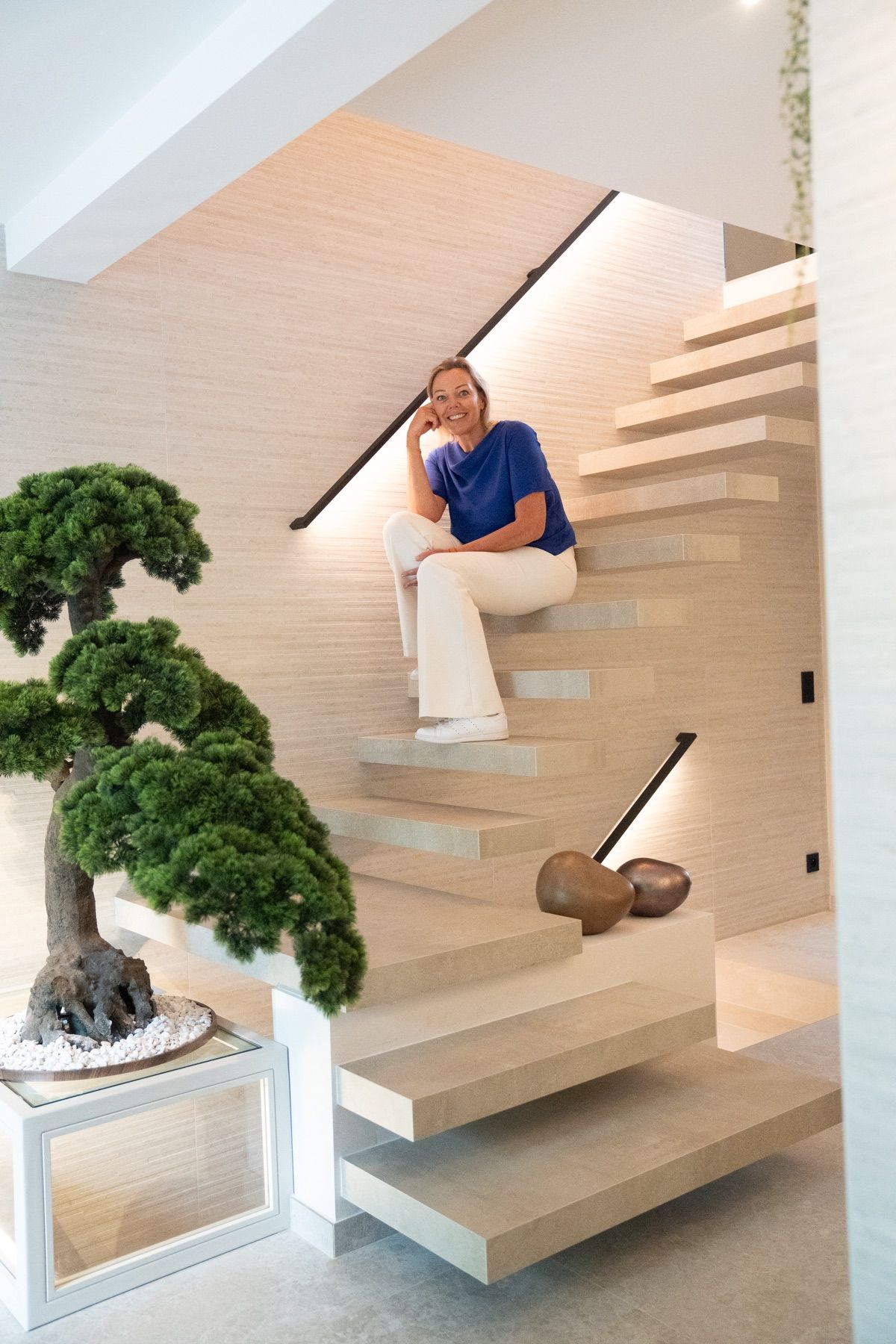 Catherine, assise sur les marches d’un escalier flottant en pierre claire, le menton posé sur la main, haut bleu et pantalon écru